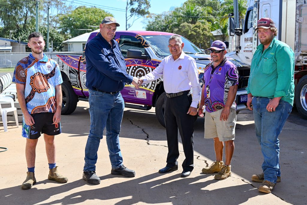 Gary White, Chairperson of Goondir Health Services, is pictured shaking hands with a man from Origin Energy as part of a solar system donation to reduce energy costs for the ACCHO. Three other men stand nearby, smiling, in front of a purple vehicle decorated with Aboriginal artwork and a truck loaded with boxed JA Solar panels. The image captures the spirit of partnership and community investment on a sunny day.
