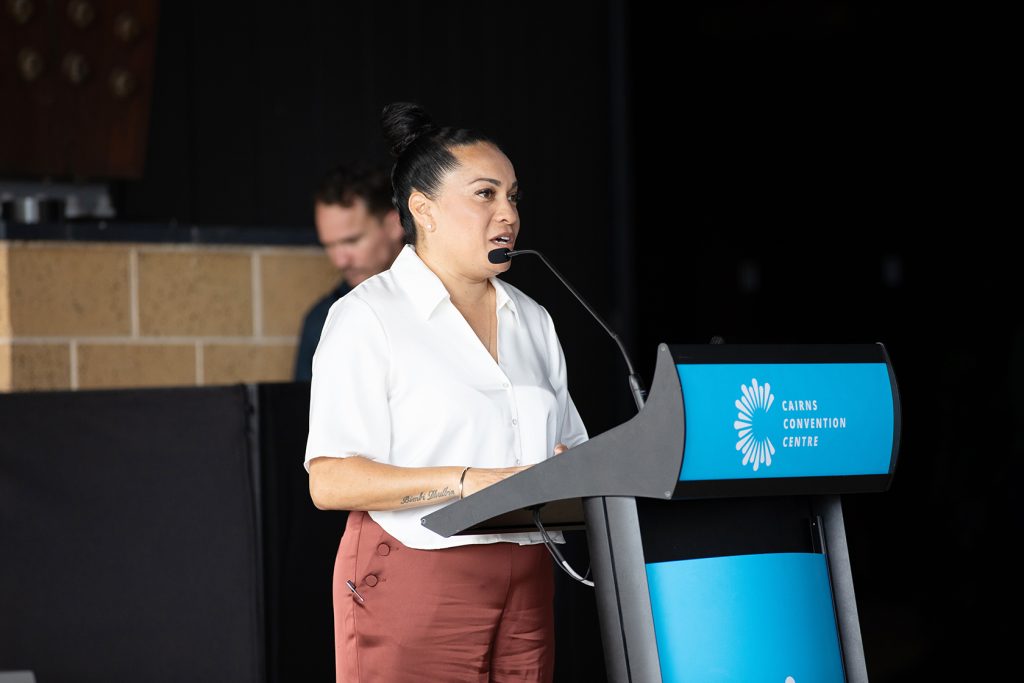 QAIHC Chairperson Dorothy Smith speaking at a podium during the October 2025 QAIHC State Members Conference at the Cairns Convention Centre. She is wearing a white button-up blouse and rust-colored trousers, addressing the audience with a headset microphone. The branded podium features the Cairns Convention Centre logo in blue and white.