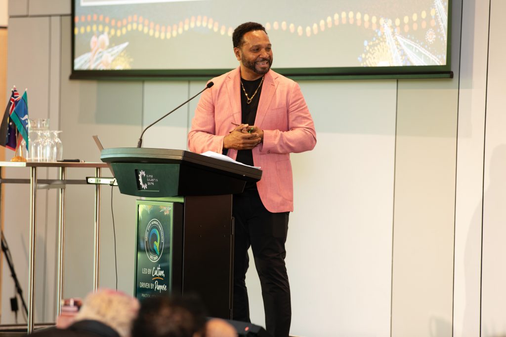 MC Dion Devow speaking at the podium during the QAIHC State Members Conference, held at the Cairns Convention Centre. He is dressed in a pink blazer over a black shirt and pants, wearing a gold necklace, and appears engaged with the audience while smiling. The lectern features conference branding with the slogan “Led by Culture, Driven by Purpose,” alongside Indigenous artwork elements. Behind him, a large presentation screen displays a dot painting-inspired design, and to the side are glass water jugs and the Australian and Torres Strait Islander flags.