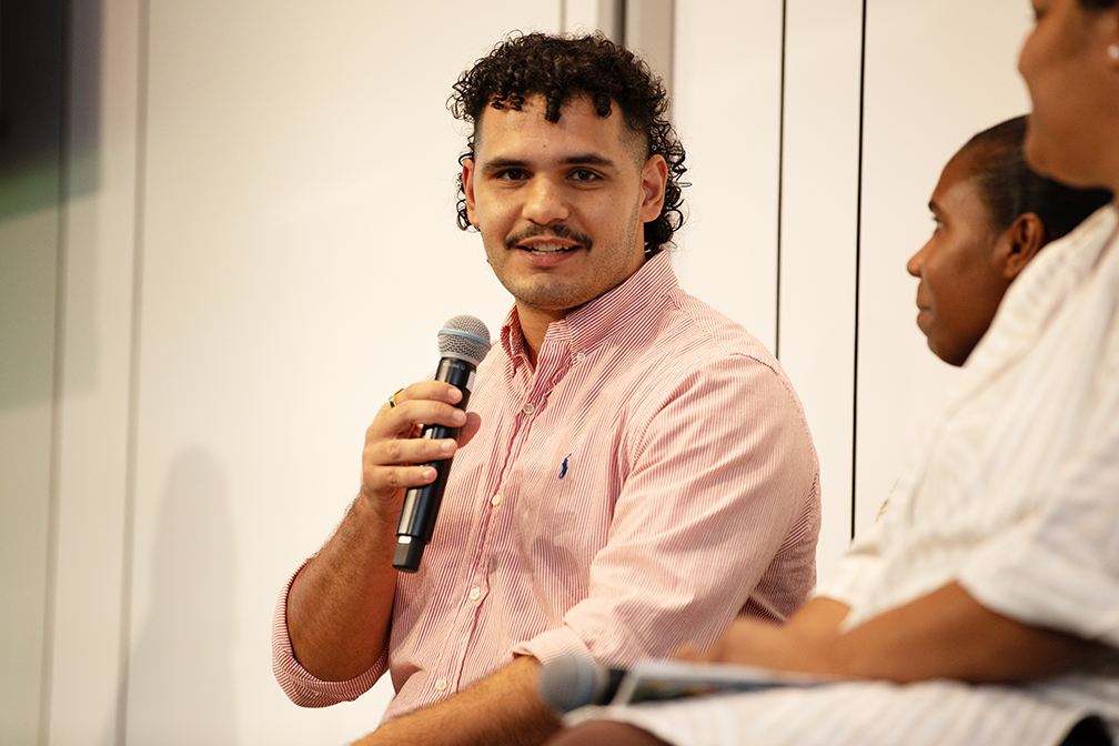 Wuchopperen board member Daniel Rosendale speaking into a microphone during a panel session at the QAIHC State Members Conference. He is wearing a pink and white striped button-up shirt with the Ralph Lauren logo and is seated among fellow panelists. Daniel is engaged with the audience, looking confidently toward the camera. The setting appears to be a formal discussion or Q&A session, with a white panel wall backdrop.