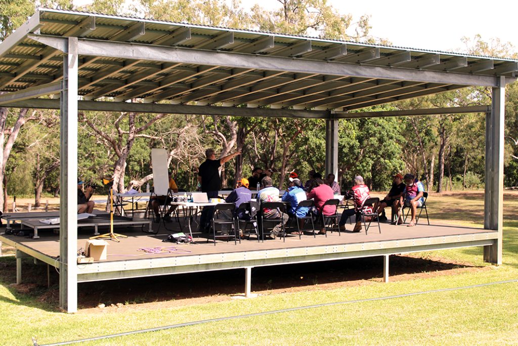 A group of men seated in work group under a large open-sided shelter during an on-Country Men's Camp. The session is taking place in a natural bushland setting, with trees and open grass visible in the background. One man stands and gestures as he speaks, while others listen and engage in the culturally significant practice of storytelling and sharing knowledge. Tables, notebooks, and a whiteboard are set up.