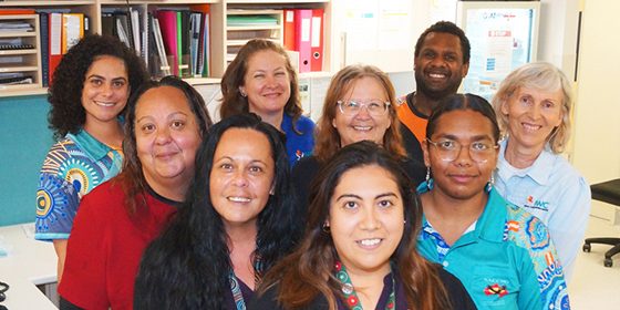 Group photo of some of the team members from the Indigenous Wellbeing Centre (IWC) in Bundaberg, standing together in a clinical office setting. The group includes a mix of Aboriginal and Torres Strait Islander and non-Indigenous staff, wearing a variety of uniforms including shirts with vibrant Indigenous artwork. They are smiling and gathered near desks, folders, and computers, reflecting a collaborative healthcare environment.