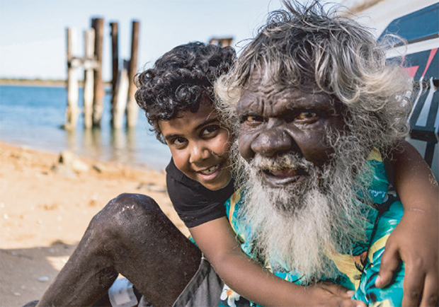 A heartwarming image of an Aboriginal Elder and a young boy smiling together on a sandy beach. The Elder has a long white beard and curly grey hair, wearing a brightly coloured shirt, while the boy hugs him from behind with a joyful expression. Wooden pylons and calm blue water are visible in the background, suggesting a coastal or remote community setting. The photo captures intergenerational connection, cultural pride, and community spirit.