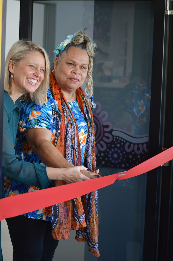 Member for Barron River, Bree James MP, and Wuchopperen Chairperson Sarah Addo cutting a ceremonial red ribbon at the official opening of the Jalbu Health Hub, a culturally safe women and girls’ health service. The pair stands in front of a glass entrance door decorated with vibrant Aboriginal artwork, symbolising community connection and cultural identity. Bree James is smiling brightly, while Sarah Addo wears a floral dress with a scarf and flower headpiece, holding ceremonial scissors